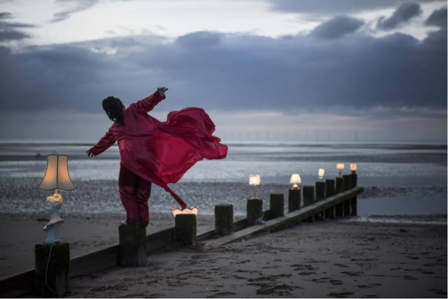 june stands on wave breakers on a beach, red gown flowing behind her in the wind, she stretches her arms out as if to fly