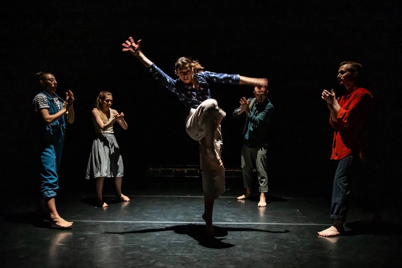 dancers clapping whilst female dancer jumps on a stage