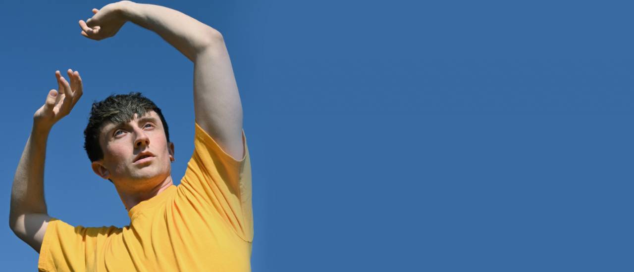 Osian in a yellow shirt lifts their hands against a blue sky