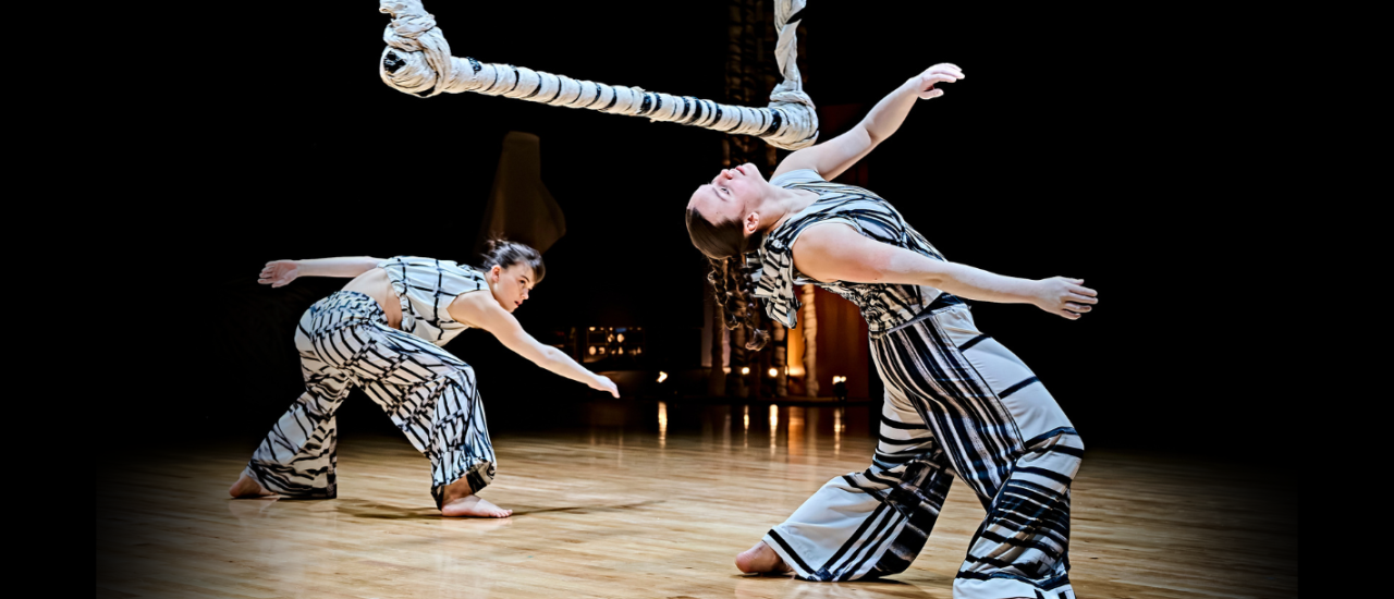 two dancers in white costumes printed with charcoal sketched lines, they bend backwards and forwards under a paper sculpture that looks like a gaint swinging bar