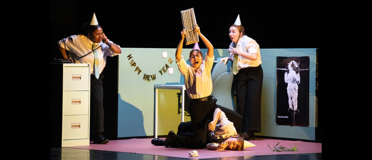 dancers in party hats and shirts and ties stand around an office looking scared, one raises a keyboard above another who is curled into a ball, the one with the keyboard is ready to strike them