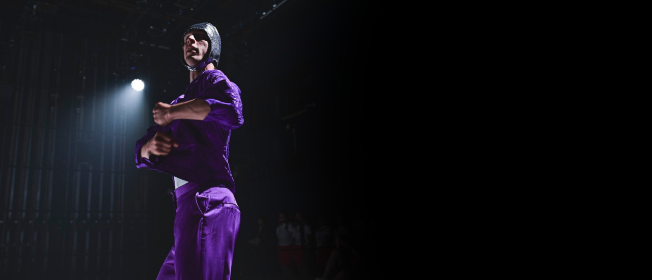 dancer in purple satin suit and glitter swim cap under dramatic lighting holds arms infront of their chest, hands balled into fists as though ready for action