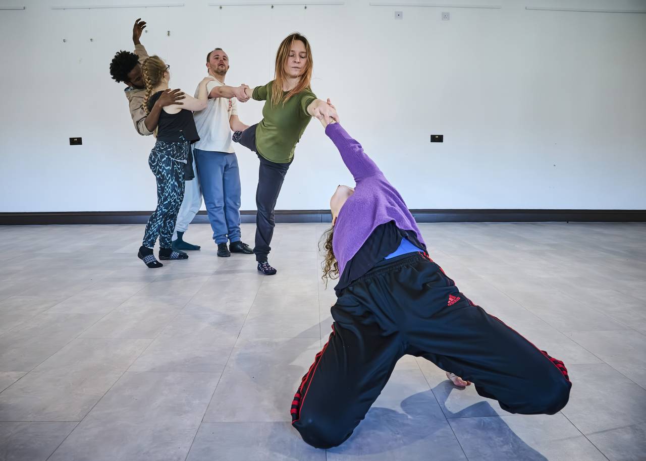 rehearsal photo, one dancer in purple stretches backwards over their own head raching for others in the back