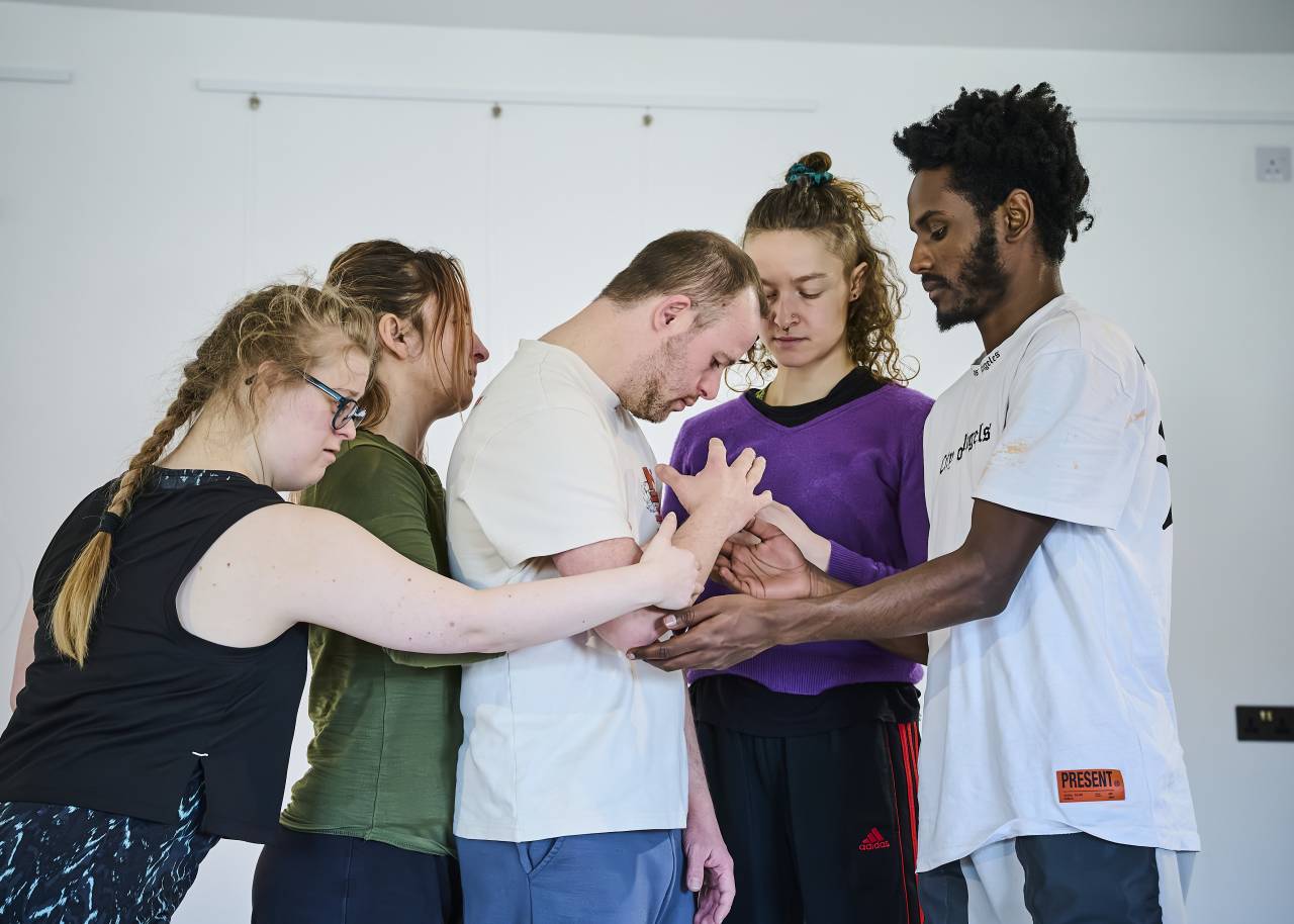four dancers huddle around another, all holdig one another tenderly