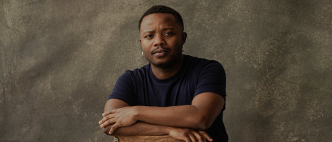 Bakani Pick-Up has short cropped hair and sits backwards on a wooden chair with a serious expression