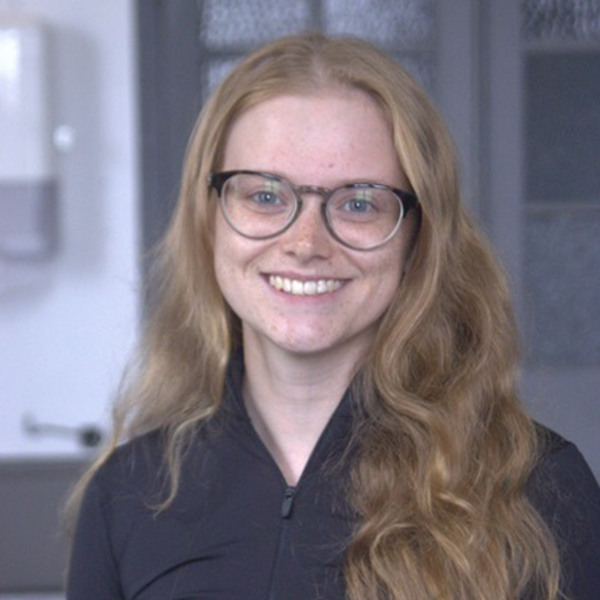 Eluned has long wavey blonde hair, dark rimmed glasses and freckles with a warm smile and wears a smart black shirt