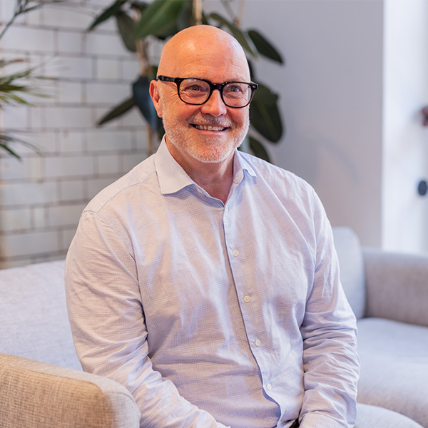 Hefin has a warm smile that crinkles his eyes. black rimmed glasses and a smart shirt, he has no hair but sports a smart white stubble beard