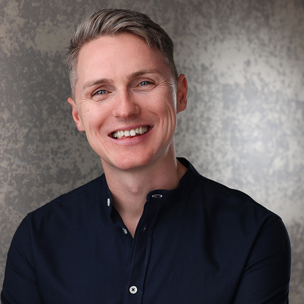 Jamie has short smart blonde hair swept back, sparkling blue eyes and a wide smile, he wears a black collared shirt